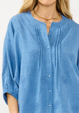 [Color: Chambray Blue] A detail image of a blonde model wearing a relaxed boho blouse with embroidery, a button front, pintuck details, and volume sleeves. 