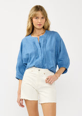 [Color: Chambray Blue] A front facing image of a blonde model wearing a relaxed boho blouse with embroidery, a button front, pintuck details, and volume sleeves. 