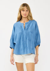 [Color: Chambray Blue] A front facing image of a blonde model wearing a relaxed boho blouse with embroidery, a button front, pintuck details, and volume sleeves. 