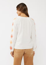 [Color: White/Coral] A back facing image of a brunette model wearing a bohemian cotton white blouse with embroidered details. Featuring a split v-neckline, long sleeves, and button wrist closure.  