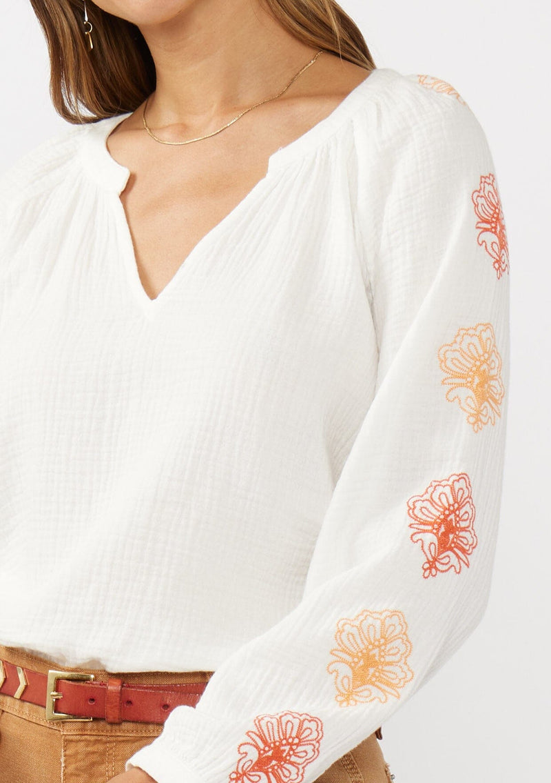 [Color: White/Coral] A detail image of a brunette model wearing a bohemian cotton white blouse with embroidered details. Featuring a split v-neckline, long sleeves, and button wrist closure.  