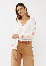[Color: White/Coral] A front facing image of a brunette model wearing a bohemian cotton white blouse with embroidered details. Featuring a split v-neckline, long sleeves, and button wrist closure.  
