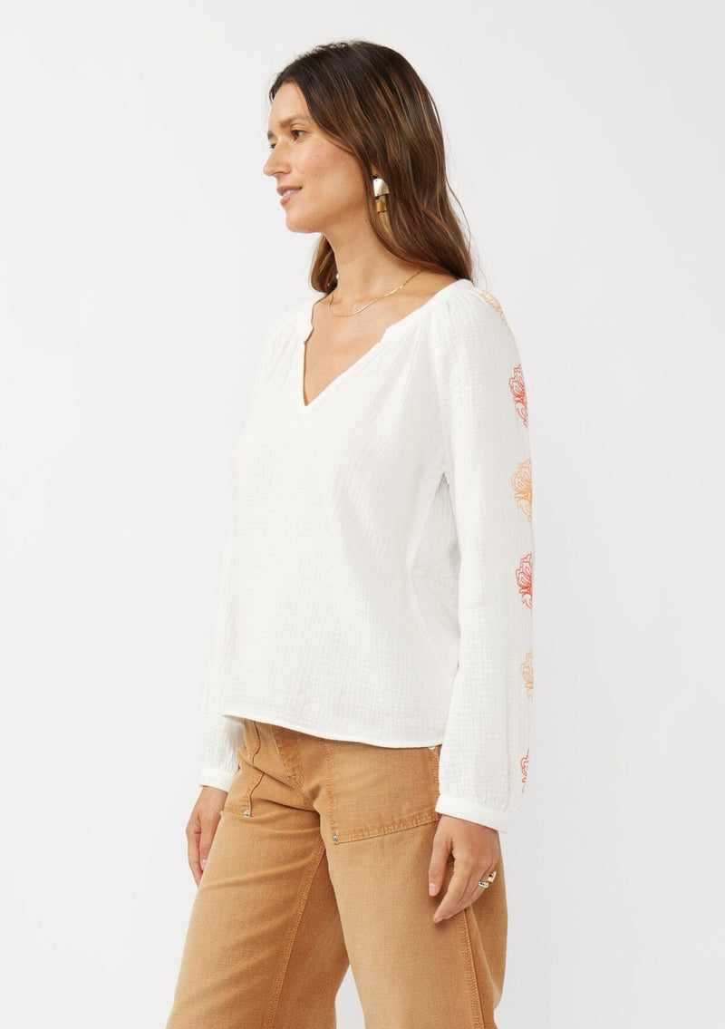 [Color: White/Coral] A side facing image of a brunette model wearing a bohemian cotton white blouse with embroidered details. Featuring a split v-neckline, long sleeves, and button wrist closure.  