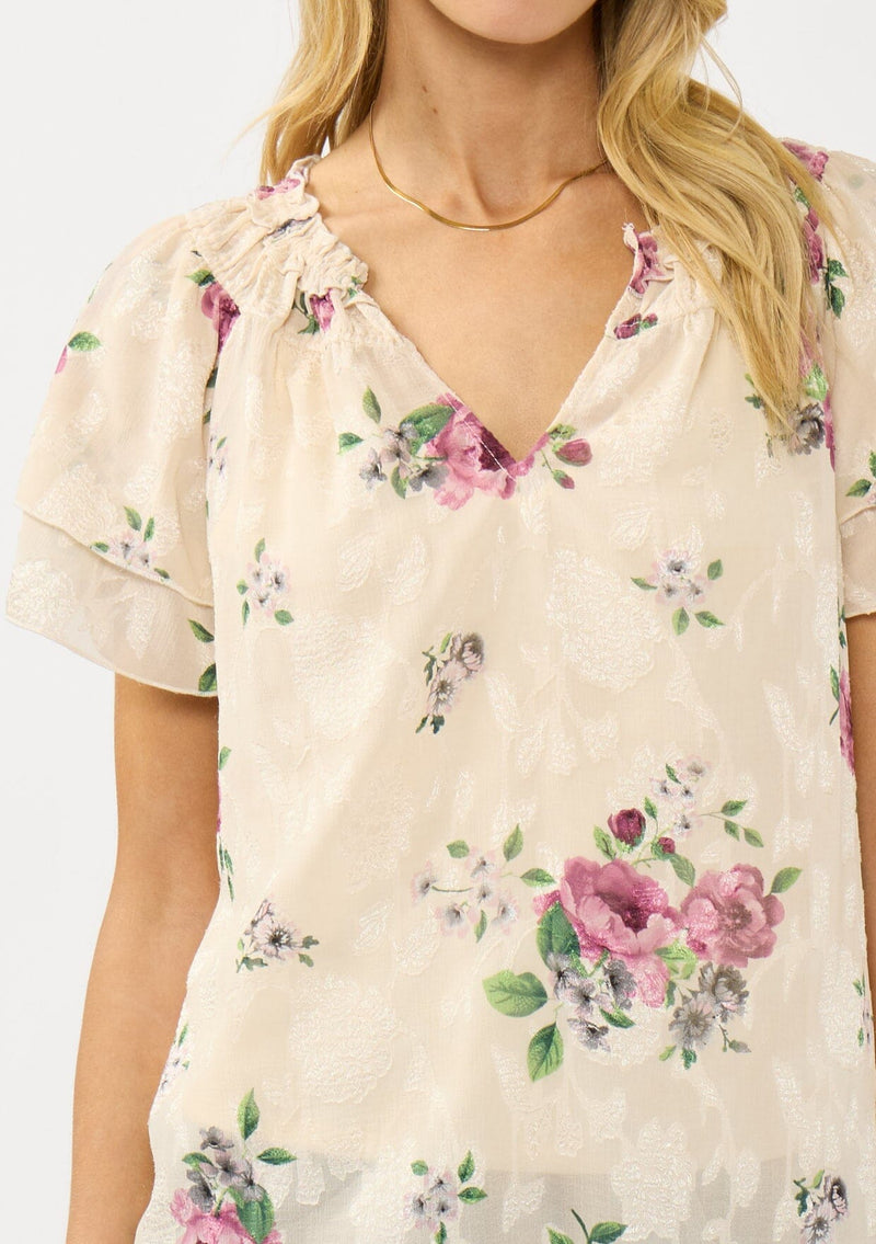 [Color: Natural/Dusty Rose] A front facing image of a blonde model wearing a pretty top with jacquard floral details and a subtle sheen. Featuring a split v-neckline, short flutter sleeves, and a ruffled neckline. 