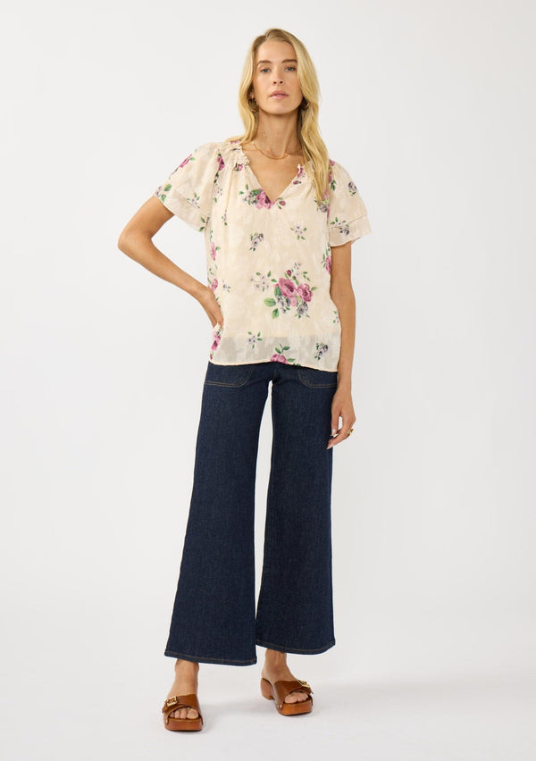 [Color: Natural/Dusty Rose] A front facing image of a blonde model wearing a pretty top with jacquard floral details and a subtle sheen. Featuring a split v-neckline, short flutter sleeves, and a ruffled neckline. 