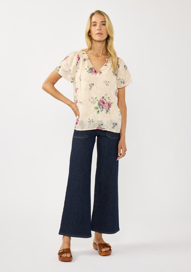 [Color: Natural/Dusty Rose] A front facing image of a blonde model wearing a pretty top with jacquard floral details and a subtle sheen. Featuring a split v-neckline, short flutter sleeves, and a ruffled neckline. 