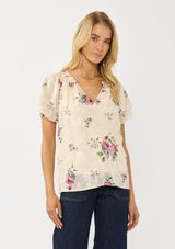 [Color: Natural/Dusty Rose] A front facing image of a blonde model wearing a pretty top with jacquard floral details and a subtle sheen. Featuring a split v-neckline, short flutter sleeves, and a ruffled neckline. 