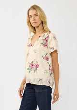 [Color: Natural/Dusty Rose] A side facing image of a blonde model wearing a pretty top with jacquard floral details and a subtle sheen. Featuring a split v-neckline, short flutter sleeves, and a ruffled neckline. 