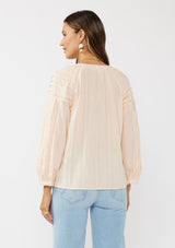 [Color: Peach] A back facing image of a brunette model wearing a light pink blouse with long raglan sleeves, a split v-neckline with button detail, lace inserts, and pintuck details. 