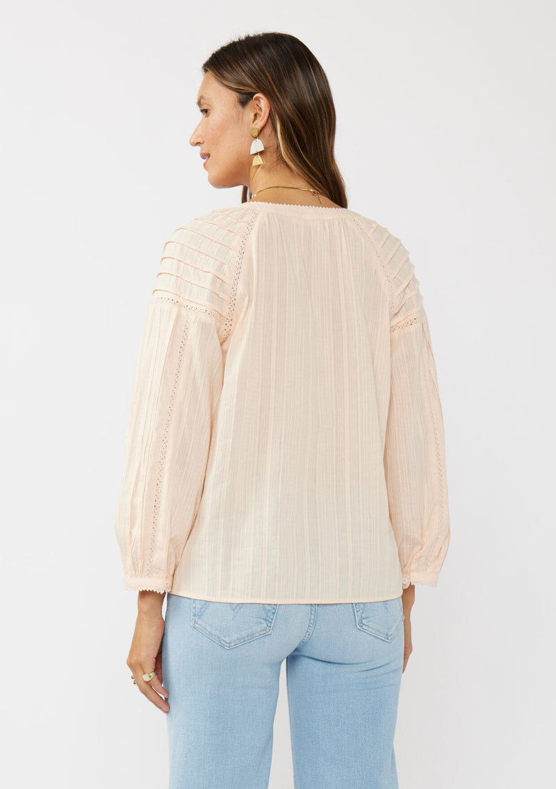 [Color: Peach] A back facing image of a brunette model wearing a light pink blouse with long raglan sleeves, a split v-neckline with button detail, lace inserts, and pintuck details. 
