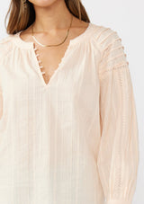 [Color: Peach] A detail image of a brunette model wearing a light pink blouse with long raglan sleeves, a split v-neckline with button detail, lace inserts, and pintuck details. 