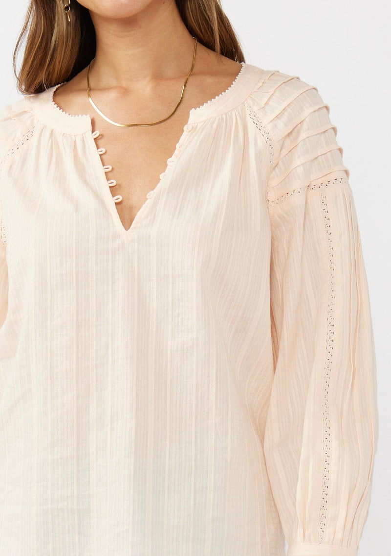 [Color: Peach] A detail image of a brunette model wearing a light pink blouse with long raglan sleeves, a split v-neckline with button detail, lace inserts, and pintuck details. 