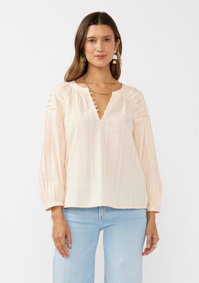 [Color: Peach] A front facing image of a brunette model wearing a light pink blouse with long raglan sleeves, a split v-neckline with button detail, lace inserts, and pintuck details. 