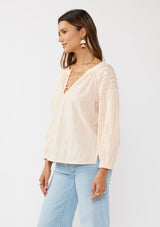 [Color: Peach] A side facing image of a brunette model wearing a light pink blouse with long raglan sleeves, a split v-neckline with button detail, lace inserts, and pintuck details. 