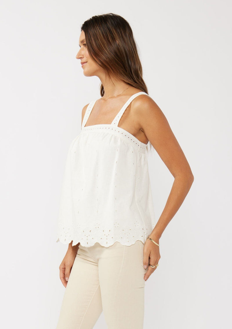 [Color: White] A side facing image of a blonde model wearing a white cotton eyelet tank top with adjustable straps and a scalloped hem. 