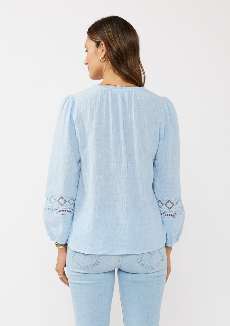 [Color: Chambray Blue] A back facing image of a brunette model wearing a light blue cotton slub blouse with long sleeves, lace details, and a split v-neckline. A versatile boho blouse perfect for the spring season. 