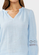 [Color: Chambray Blue] A detail image of a brunette model wearing a light blue cotton slub blouse with long sleeves, lace details, and a split v-neckline. A versatile boho blouse perfect for the spring season. 