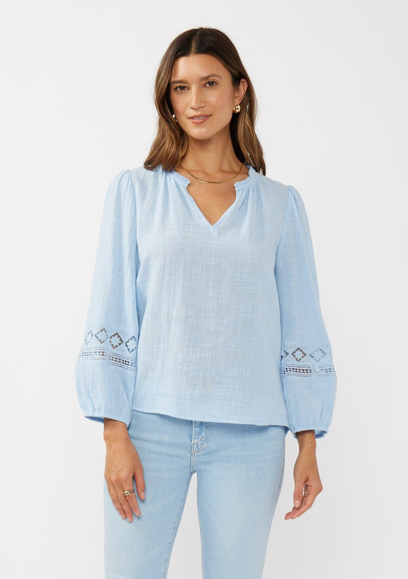 [Color: Chambray Blue] A front facing image of a brunette model wearing a light blue cotton slub blouse with long sleeves, lace details, and a split v-neckline. A versatile boho blouse perfect for the spring season. 