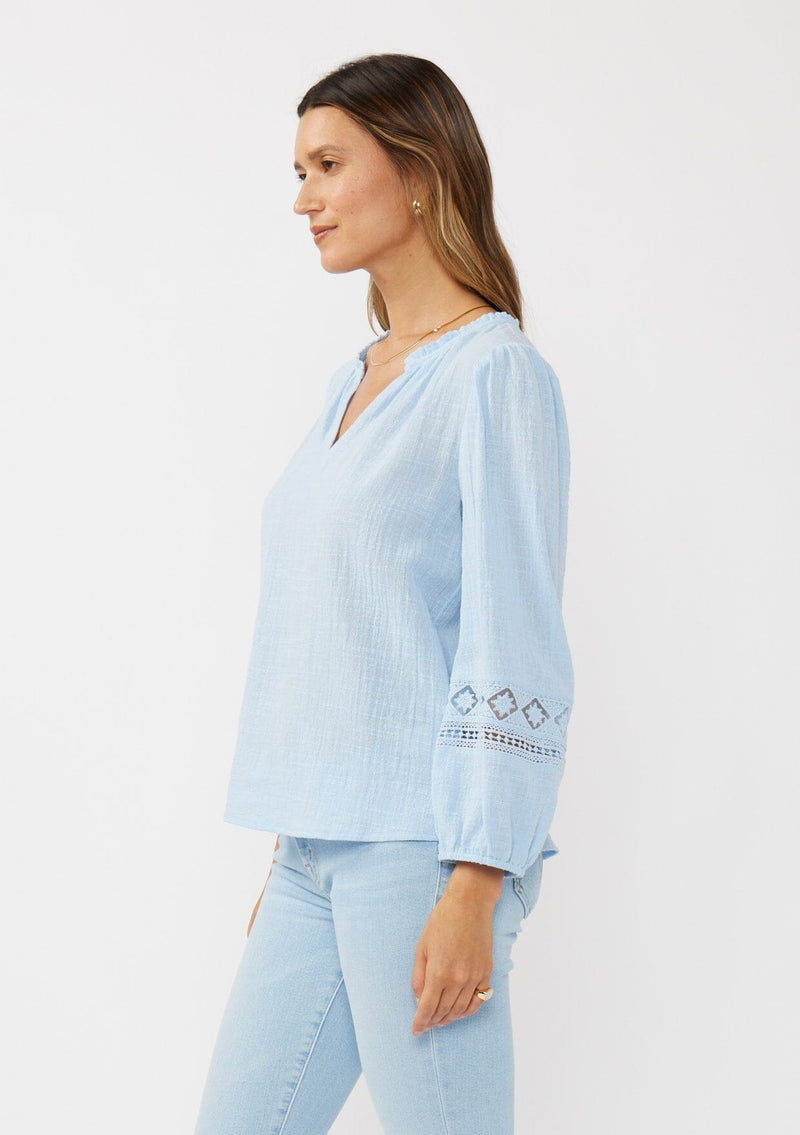 [Color: Chambray Blue] A side facing image of a brunette model wearing a light blue cotton slub blouse with long sleeves, lace details, and a split v-neckline. A versatile boho blouse perfect for the spring season. 