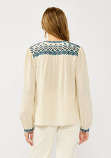[Color: Vanilla/Dusty Turquoise] A back facing image of a blonde model wearing a cream cotton slub blouse with blue embroidered details, button front, a round neckline, and long sleeves. 