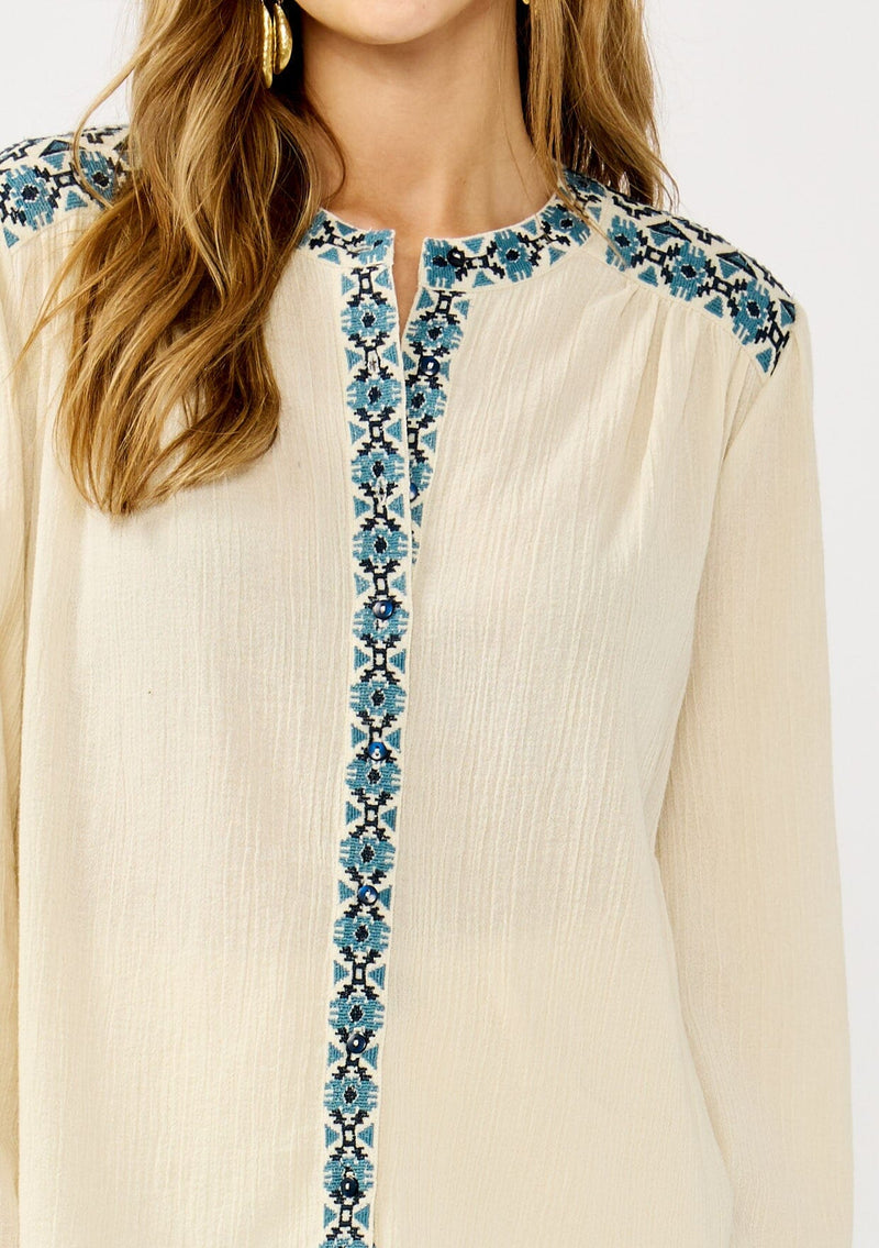 [Color: Vanilla/Dusty Turquoise] A detail image of a blonde model wearing a cream cotton slub blouse with blue embroidered details, button front, a round neckline, and long sleeves. 