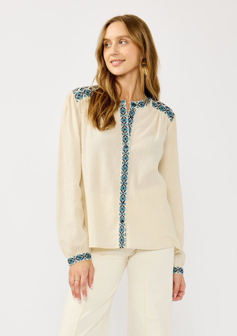 [Color: Vanilla/Dusty Turquoise] A front facing image of a blonde model wearing a cream cotton slub blouse with blue embroidered details, button front, a round neckline, and long sleeves. 