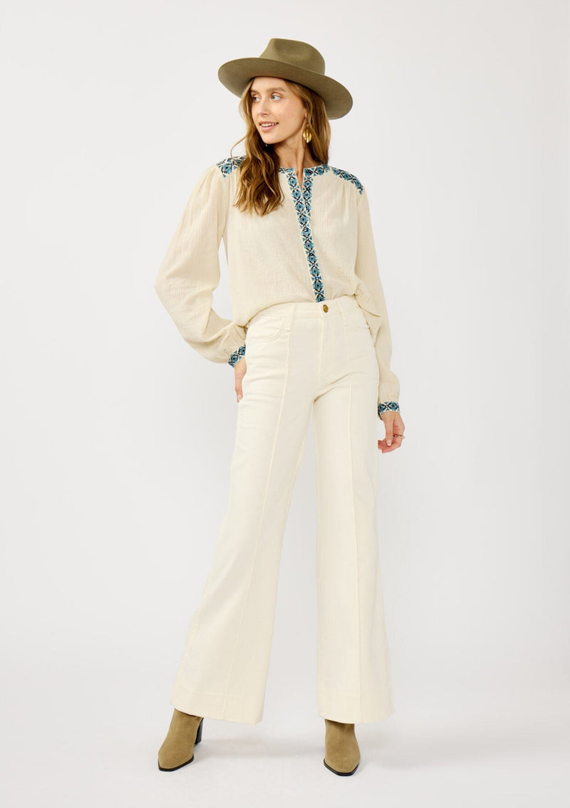 [Color: Vanilla/Dusty Turquoise] A front facing image of a blonde model wearing a cream cotton slub blouse with blue embroidered details, button front, a round neckline, and long sleeves. 
