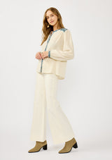 [Color: Vanilla/Dusty Turquoise] A side facing image of a blonde model wearing a cream cotton slub blouse with blue embroidered details, button front, a round neckline, and long sleeves. 