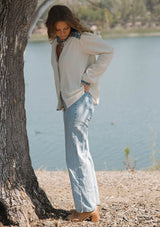 [Color: Vanilla/Dusty Turquoise] A side facing image of a blonde model wearing a cream cotton slub blouse with blue embroidered details, button front, a round neckline, and long sleeves. 