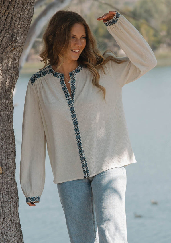 [Color: Vanilla/Dusty Turquoise] A front facing image of a blonde model wearing a cream cotton slub blouse with blue embroidered details, button front, a round neckline, and long sleeves. 