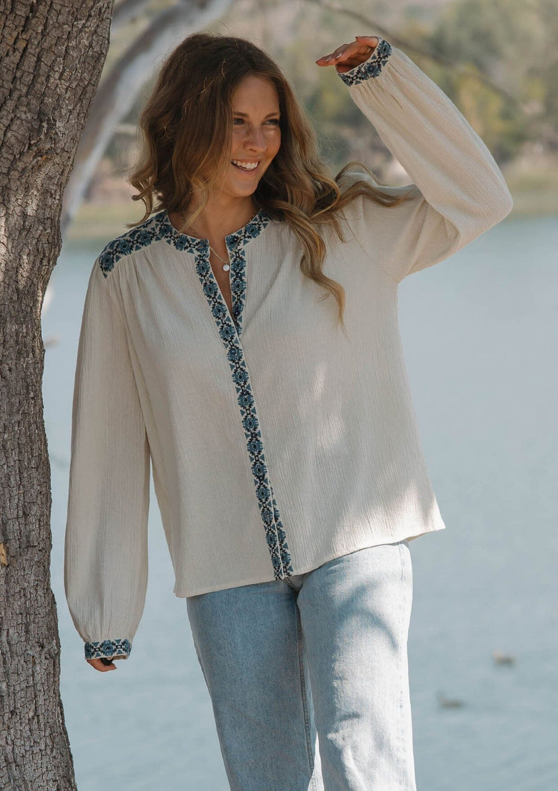 [Color: Vanilla/Dusty Turquoise] A front facing image of a blonde model wearing a cream cotton slub blouse with blue embroidered details, button front, a round neckline, and long sleeves. 