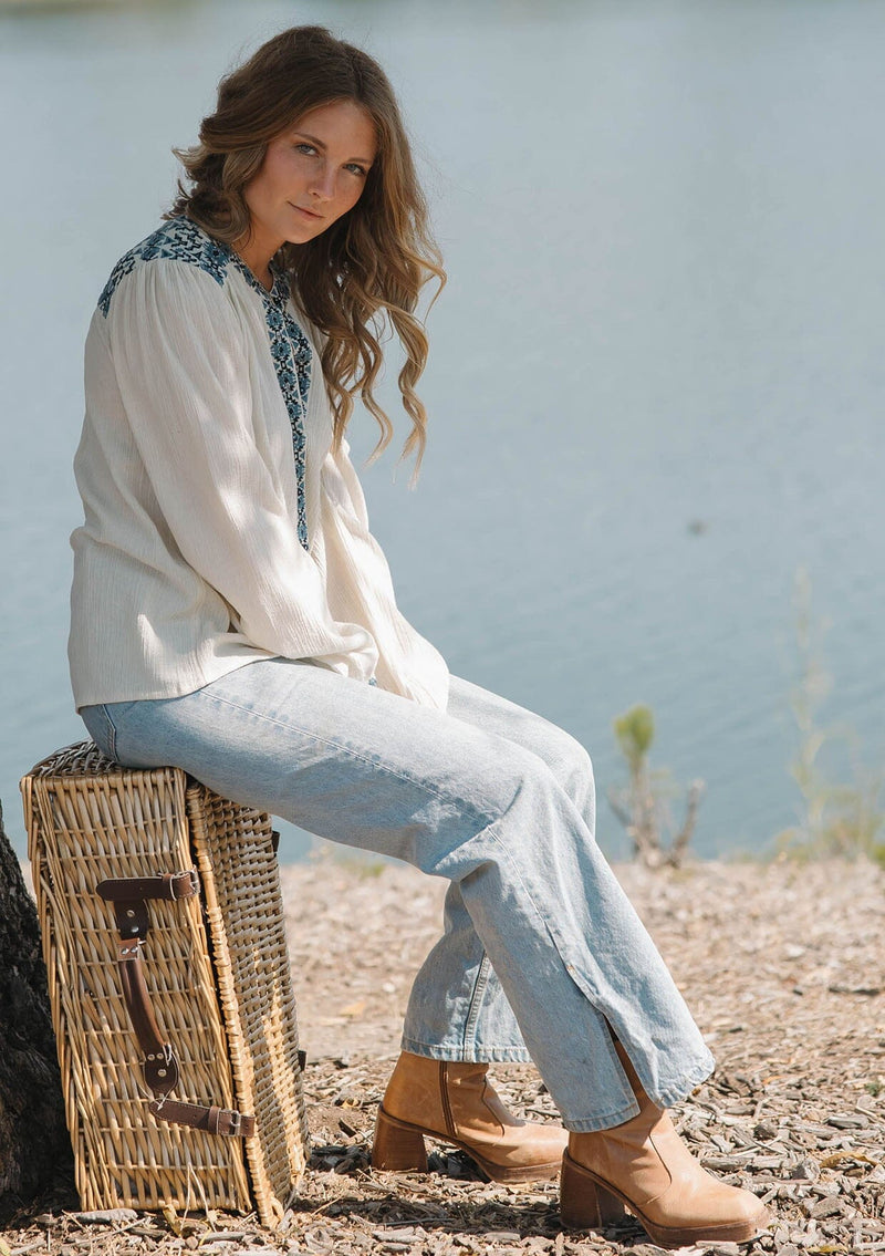 [Color: Vanilla/Dusty Turquoise] A sitting image of a blonde model wearing a cream cotton slub blouse with blue embroidered details, button front, a round neckline, and long sleeves. 