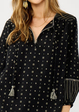 [Color: Black/Taupe] A detail image of a blonde model wearing a black and taupe cotton jacquard blouse with a split v-neckline, tassel ties, long sleeves, and blanket stitch details. A fall bohemian top. 