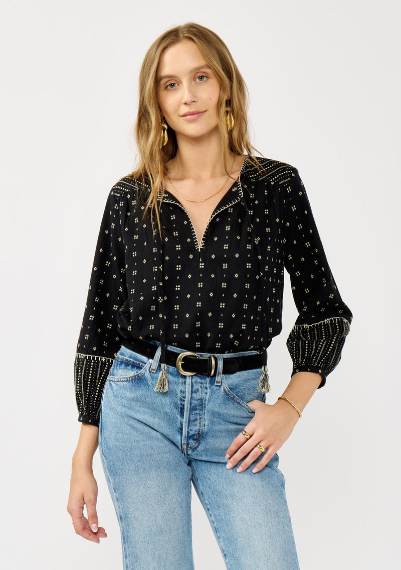 [Color: Black/Taupe] A front facing image of a blonde model wearing a black and taupe cotton jacquard blouse with a split v-neckline, tassel ties, long sleeves, and blanket stitch details. A fall bohemian top. 