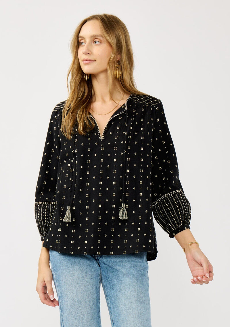 [Color: Black/Taupe] A front facing image of a blonde model wearing a black and taupe cotton jacquard blouse with a split v-neckline, tassel ties, long sleeves, and blanket stitch details. A fall bohemian top. 