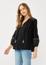 [Color: Black/Taupe] A side facing image of a blonde model wearing a black and taupe cotton jacquard blouse with a split v-neckline, tassel ties, long sleeves, and blanket stitch details. A fall bohemian top. 