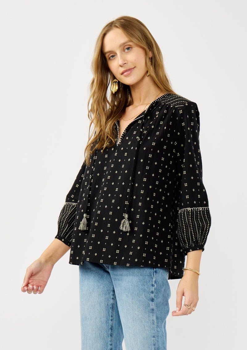 [Color: Black/Taupe] A side facing image of a blonde model wearing a black and taupe cotton jacquard blouse with a split v-neckline, tassel ties, long sleeves, and blanket stitch details. A fall bohemian top. 