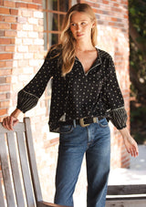 [Color: Black/Taupe] A front facing image of a blonde model wearing a black and taupe cotton jacquard blouse with a split v-neckline, tassel ties, long sleeves, and blanket stitch details. A fall bohemian top. 