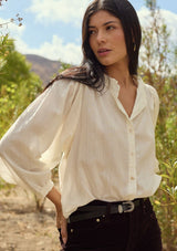 [Color: Cream] A front facing image of a brunette model outside wearing an ivory button front top with a high ruffled neckline, long sleeves, and smocked elastic cuffs. A relaxed fit fall blouse perfect for the office and special occasions. 