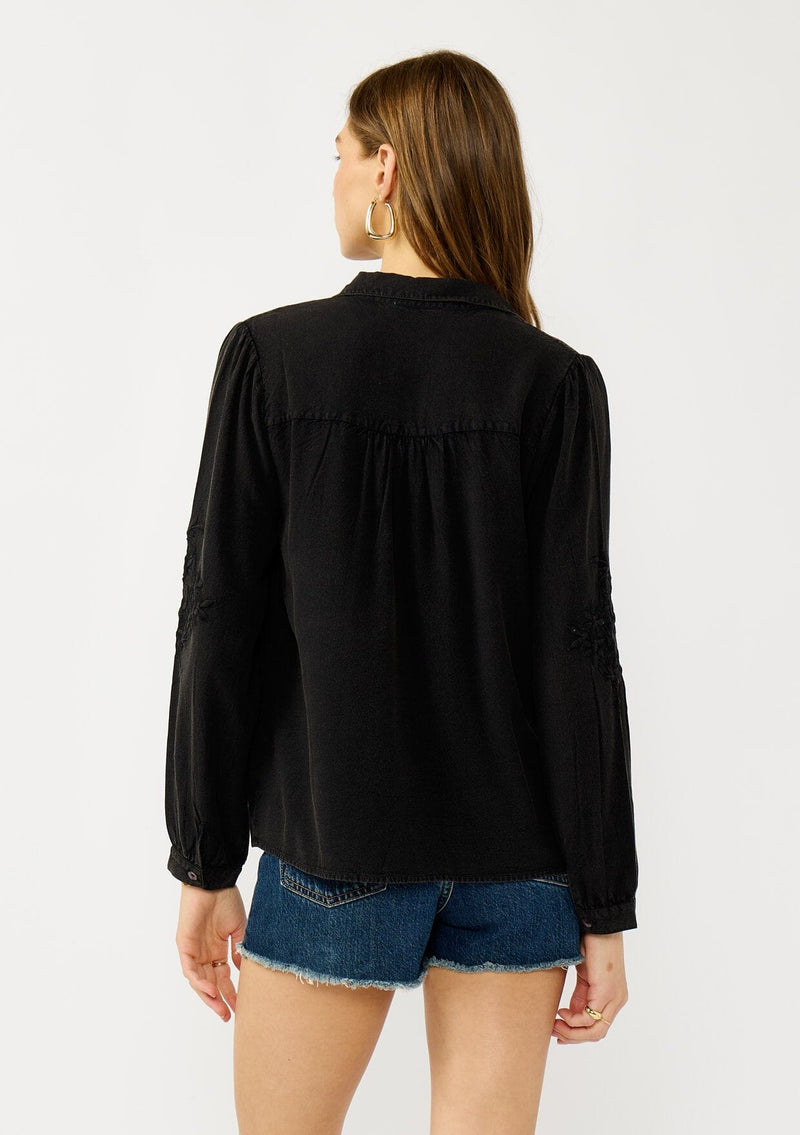 [Color: Black] A back facing image of a blonde model wearing a soft vintage wash black shirt with tonal embroidered details. Featuring a classic collar neckline, button front, and button cuff. 