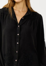 [Color: Black] A detail image of a blonde model wearing a soft vintage wash black shirt with tonal embroidered details. Featuring a classic collar neckline, button front, and button cuff. 