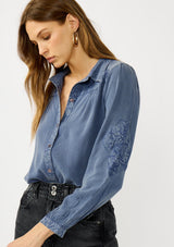 [Color: Denim Blue] A front facing image of a blonde model wearing a blue vintage wash shirt with floral embroidery, a classic collared neckline and button front. 