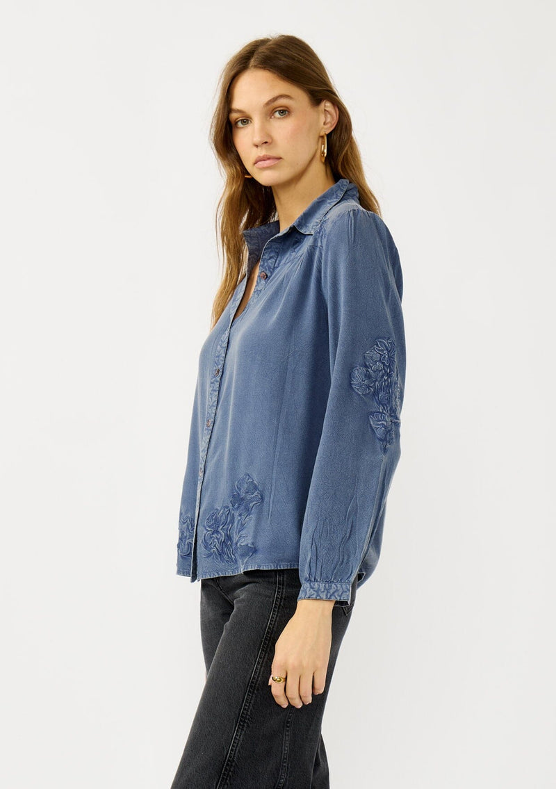 [Color: Denim Blue] A side facing image of a blonde model wearing a blue vintage wash shirt with floral embroidery, a classic collared neckline and button front. 