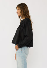 [Color: Black] A side facing image of a brunette model wearing black lightweight blouse with pintuck accents, a button front, and split v-neckline. Airy and sheer for any warm weather occasion. 