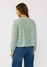 [Color: Dusty Sage] A back facing image of a brunette model wearing green lightweight blouse with pintuck accents, a button front, and split v-neckline. Airy and sheer for any warm weather occasion. 
