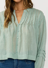 [Color: Dusty Sage] A detail image of a brunette model wearing green lightweight blouse with pintuck accents, a button front, and split v-neckline. Airy and sheer for any warm weather occasion. 