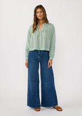 [Color: Dusty Sage] A front facing image of a brunette model wearing green lightweight blouse with pintuck accents, a button front, and split v-neckline. Airy and sheer for any warm weather occasion. 