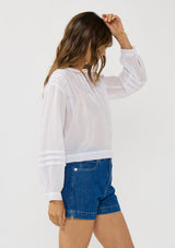 [Color: Black] A side facing image of a brunette model wearing white lightweight blouse with pintuck accents, a button front, and split v-neckline. Airy and sheer for any warm weather occasion. 