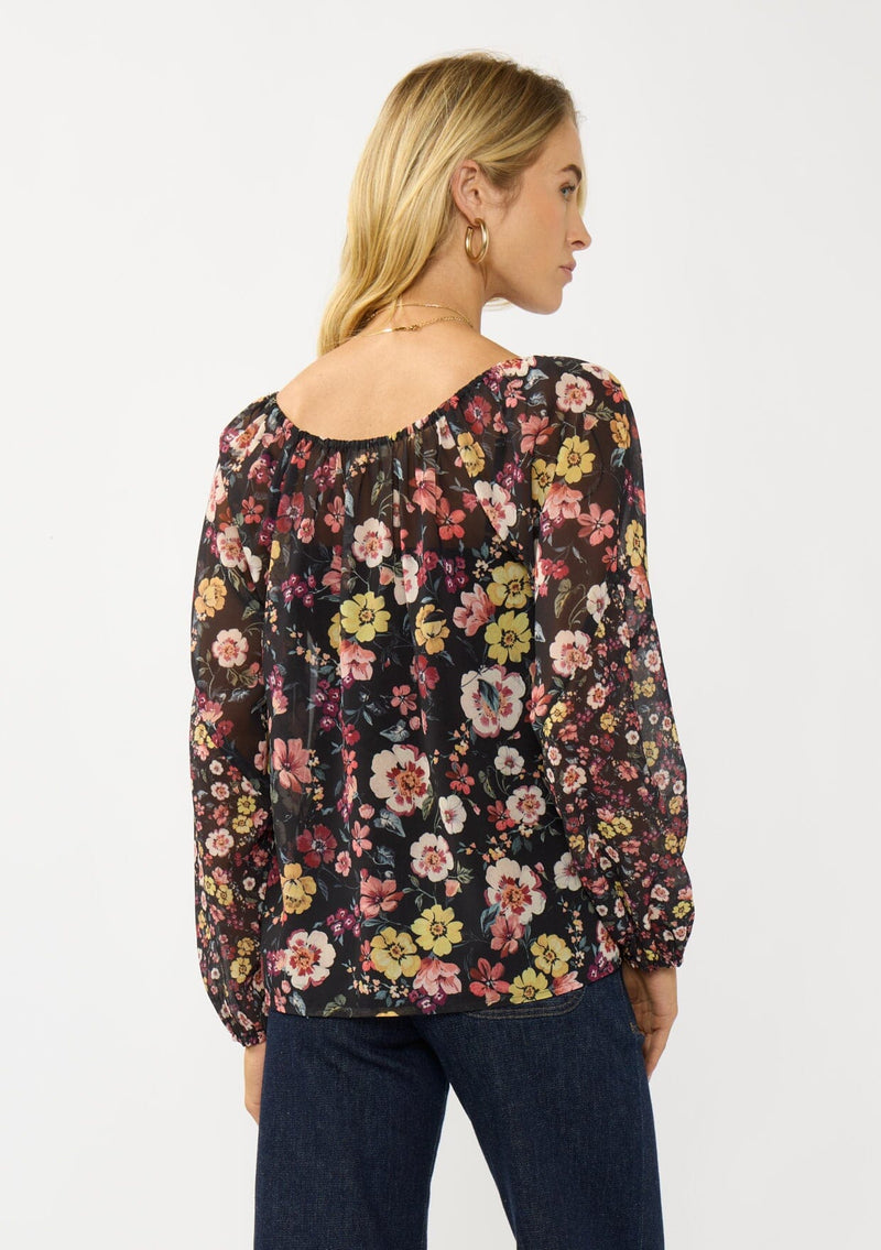 [Color: Black/Rust] A back facing image of a blonde model wearing a black floral sheer chiffon blouse with a split v-neckline with tie detail and long volume sleeves. 