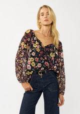 [Color: Black/Rust] A front facing image of a blonde model wearing a black floral sheer chiffon blouse with a split v-neckline with tie detail and long volume sleeves. 